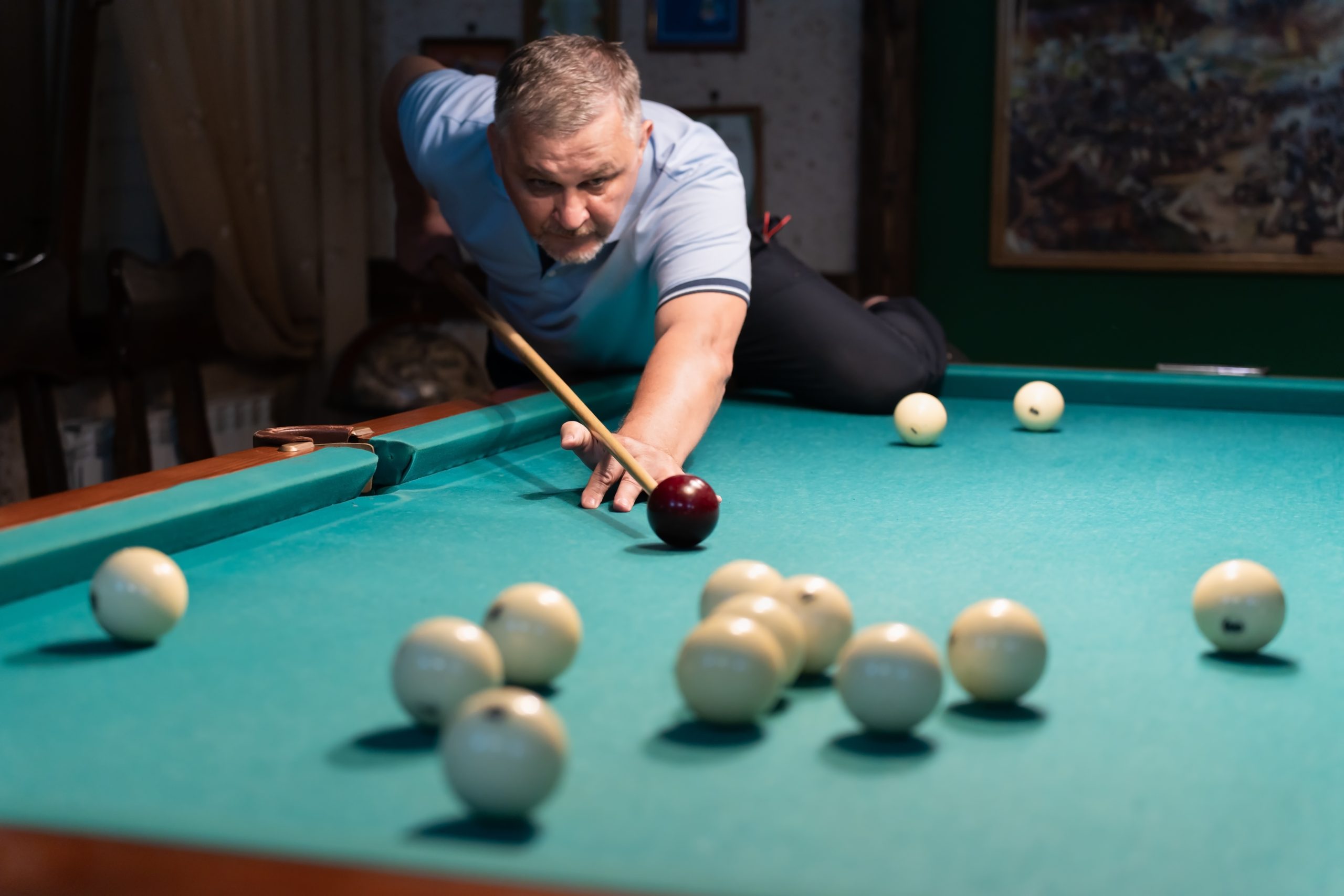 Concentrated mature man playing billiards, carefully aiming his cue stick to hit the white ball on a green billiard table in a dimly lit game room