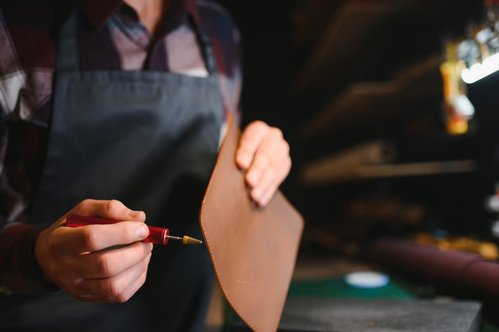 Young focused experienced master craftsman makes leather goods at his workplace